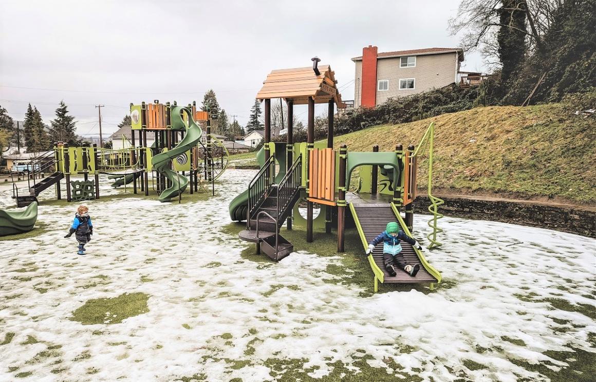 Wide view of the new playground at South Seattle’s Lakeridge Playfield in a slightly snowy day kids play on green and brown nature-themed play equipment
