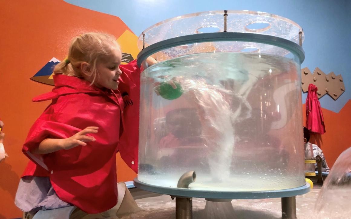 Young girl wearing plastic apron plays with a water feature at the Discovery Children’s Museum in Las Vegas, great sunny destination for Seattle area families