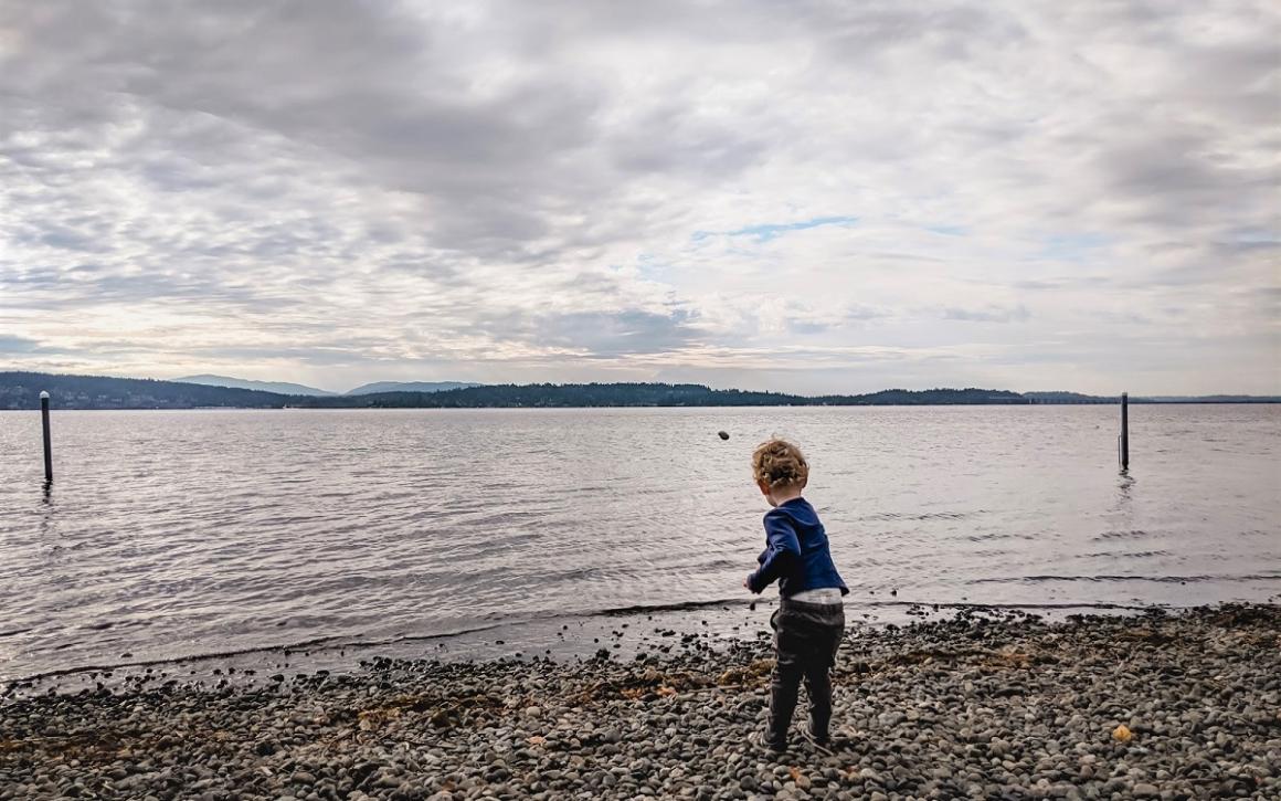 Small boy standing on the rocky shore of Lake Washington throw rocks into the water at magnuson park