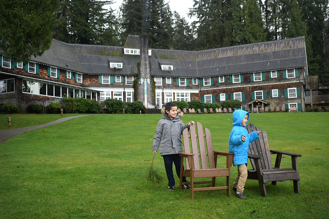 Two boys in winter jackets standing next to adirondack chairs on the lawn in front of historic Lake Quinault Lodge on Washington's Olympic Peninsula