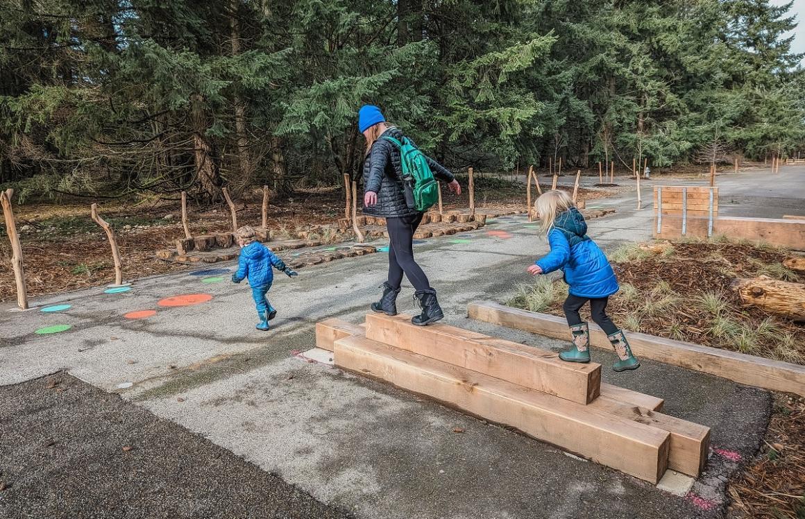 Family walking on natural balance beam made of wood among natural play elements at Pause and Play nature play area Swan Creek Park Tacoma