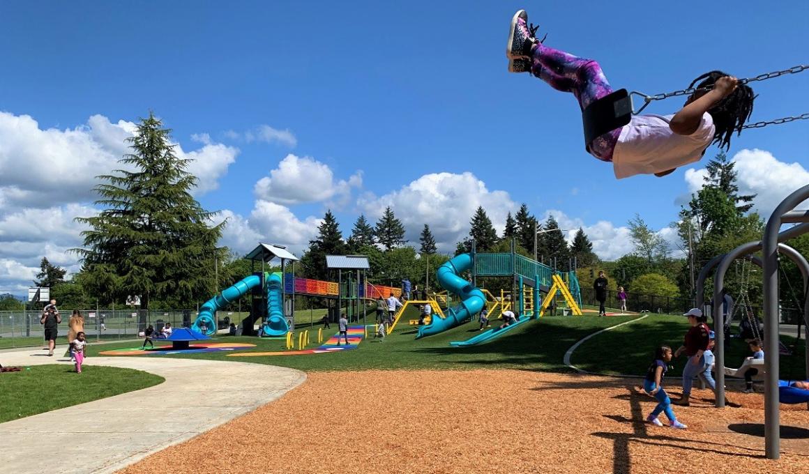 Girl high on swing in the foreground with West Fenwick Slides and Climbers lift size game in the background new playground near Seattle