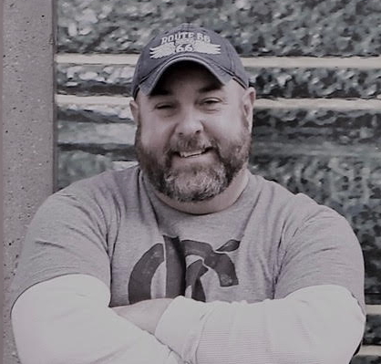 Shannon Carpenter standing in front of wall with his arms crossed smiling and wearing a baseball hat