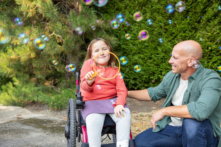 Emma in a wheelchair blowing bubbles outside with her dad