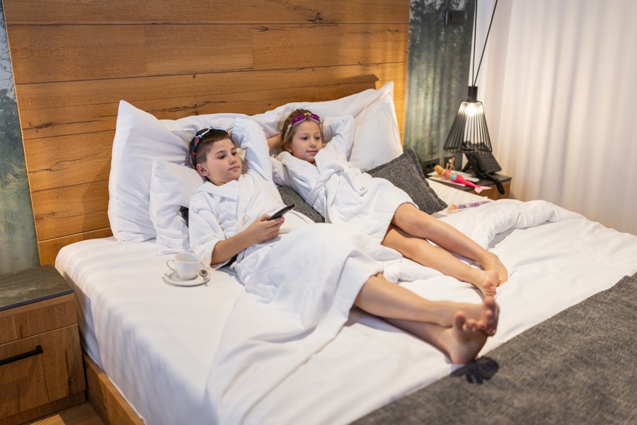 two kids relaxing on a hotel bed wearing bathrobes
