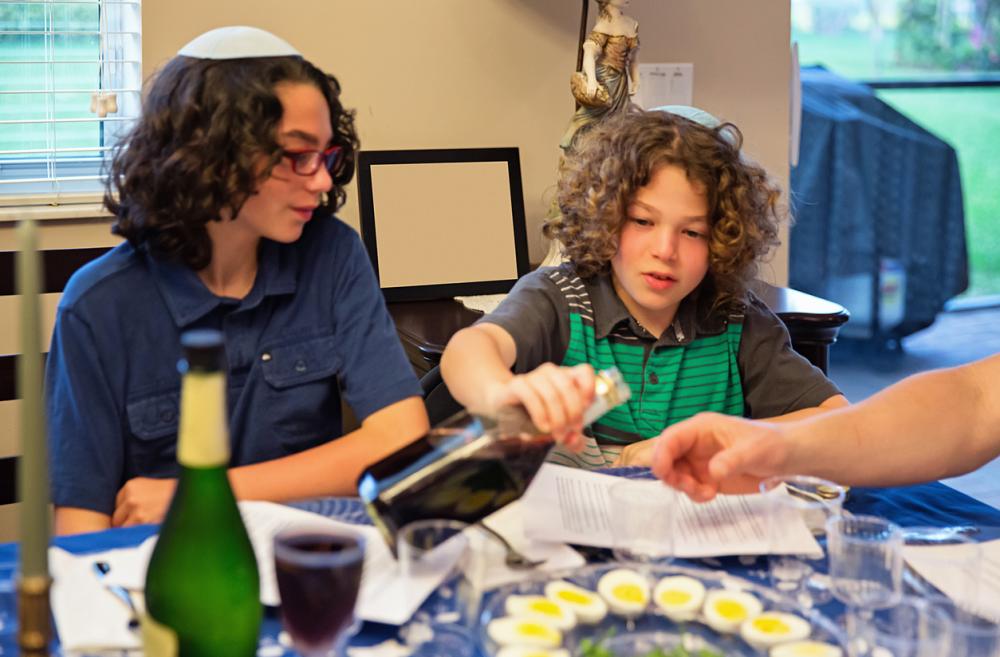 Two brothers with brown curly hair sit at the family table participating in a Passover Seder with wine and hardboiled eggs