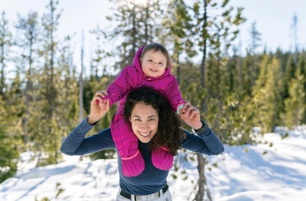 Woman holding a smiling baby on her shoulders in the woods with snow on the ground