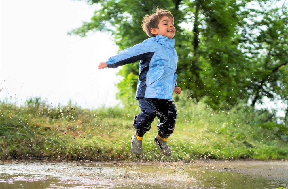 A young boy in a light blue rain jacket, pants and boots jumps gleefully in a mud puddle with green trees and shrubs behind him