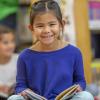 A super cute elementary school girl of Filipino ethnicity sits on the floor reading a book at her school library