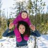Woman holding a smiling baby on her shoulders in the woods with snow on the ground