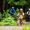 A young girl rides a bike on a bike path with her older sibling and parent following behind. They are wearing helmets and the paved path is lined with green plants indicating summer.