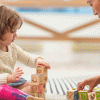 a young girl plays with wooden blocks along with an adult on a rug, safe toys