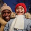 A father and daughter dressed for the cold stand in front of a decorated tree.