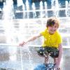 A young boy in a yellow shirt crouches down at a splash pad and enjoys the water.