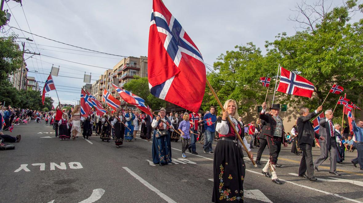 People in Norwegian traditional costumes walk in Ballard Syttende Mai parade holding Norwegian flags