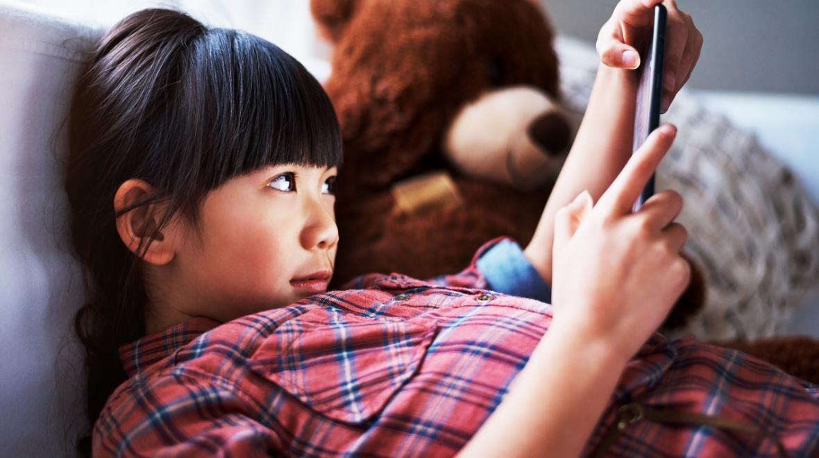 Girl at home looking at tablet sitting on couch with teddy bear beside her