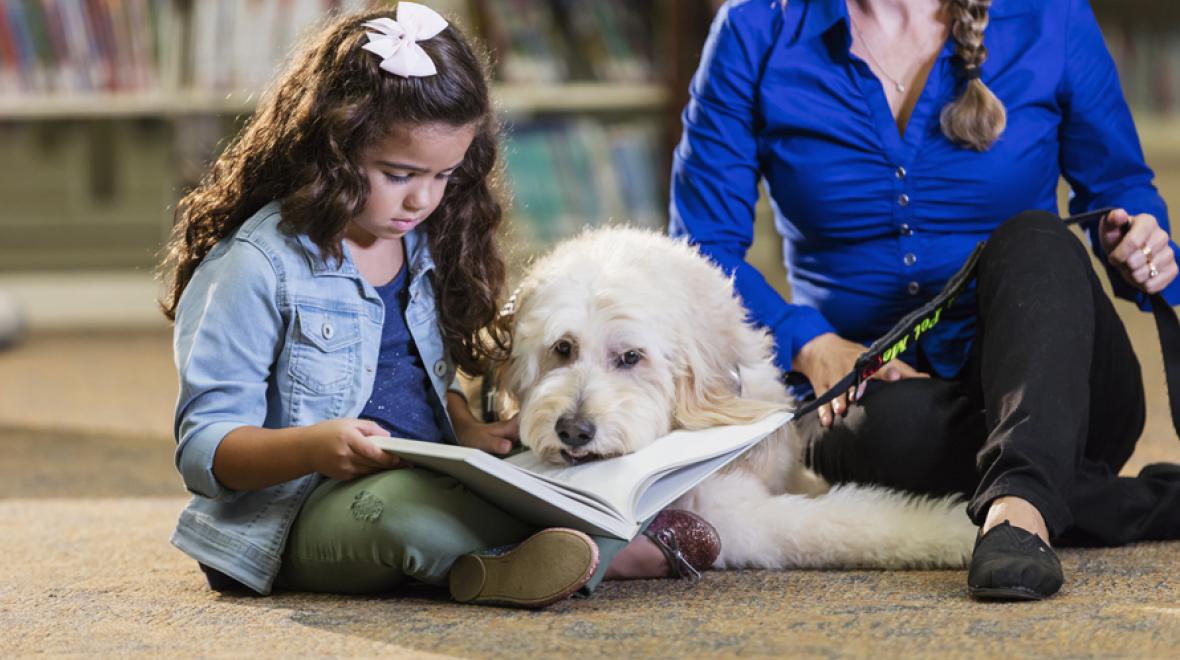 little girl reading to a dog in the library with a librarian nearby