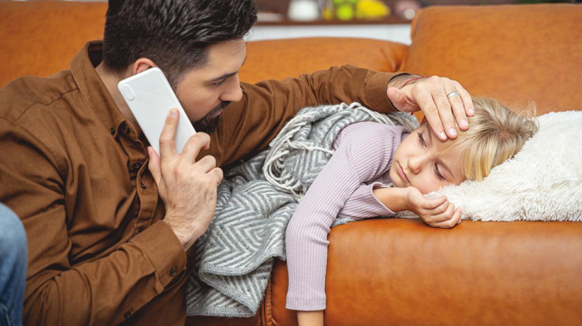 dad holding a hand to his sick daughter's forehead while holding the phone