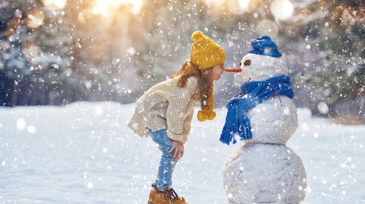 girl in a yellow hat bumping noses/carrot with a snowman wearing a blue scarf