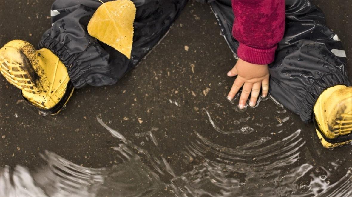 Child sitting in a rain puddle playing, wearing yellow boots and holding a yellow leaf