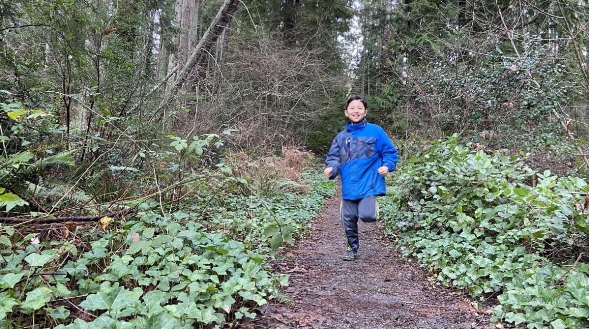 Happy kid in blue jacket running on a forested trail on Bainbridge Island near Gazzam Lake Nature Preserve