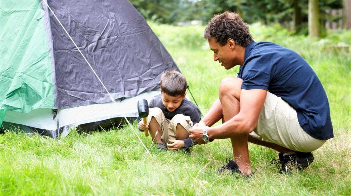 Father in shorts and a blue polo shirt with a young son together setting up a camping tent in lush green grass