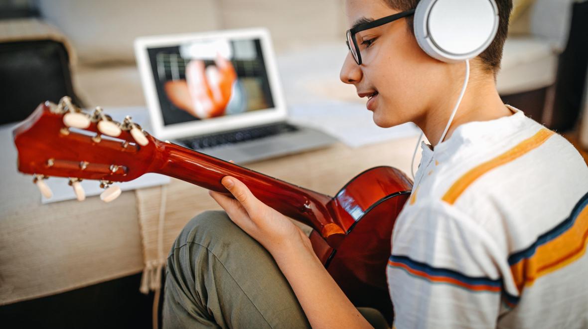 teenager sitting in front of a youtube video on a laptop holding a guitar