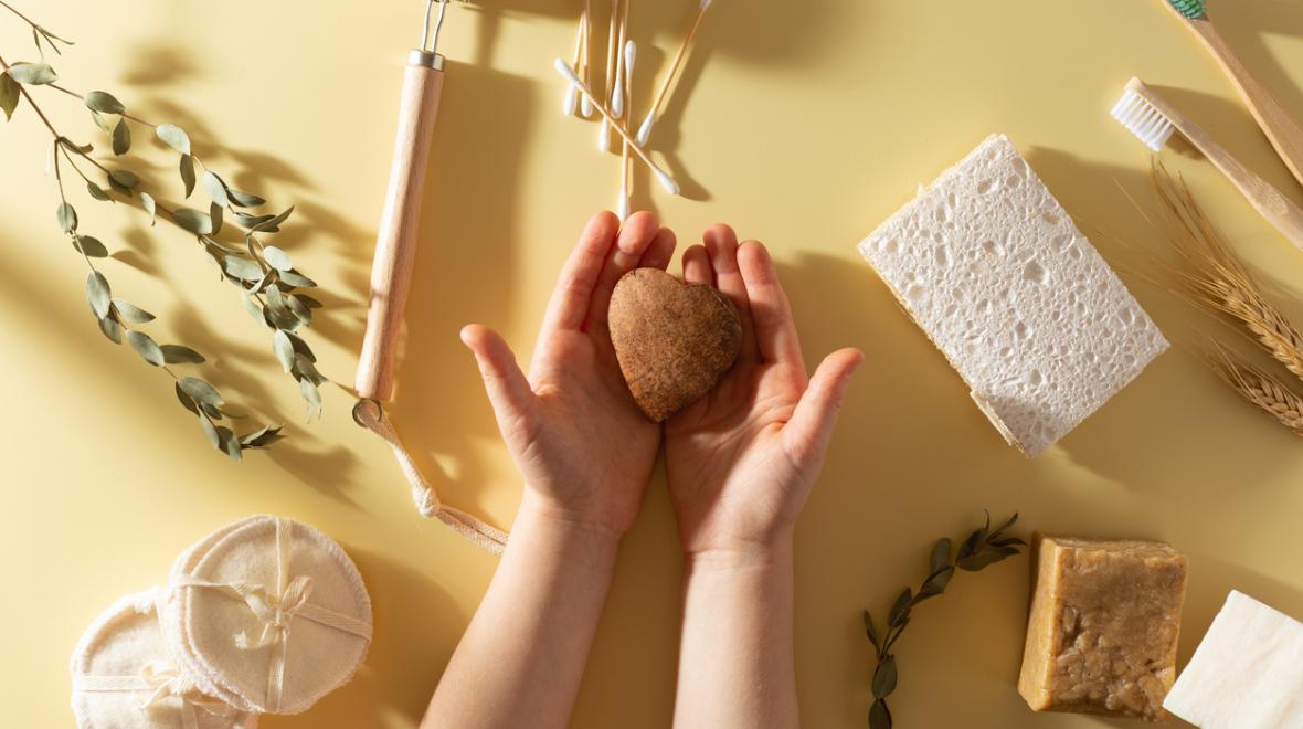 child's hands holding a heart shaped bar of organic soap surrounded by other sustainable products like a bamboo toothbrush and eco-friendly sponges