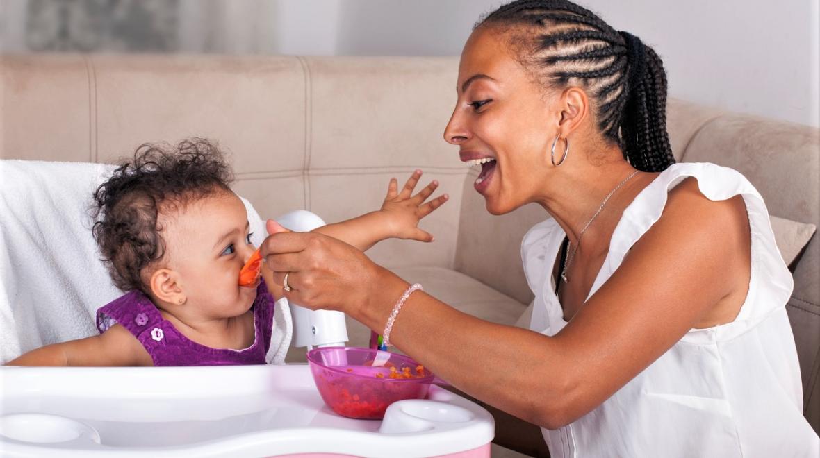 Black mom smiling and feeding baby in high chair with spoon in kitchen avoiding toxic ingredients in processed baby foods