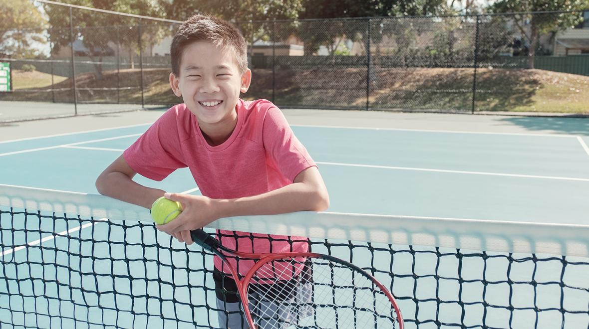 boy leaning against a tennis net holding a tennis racket