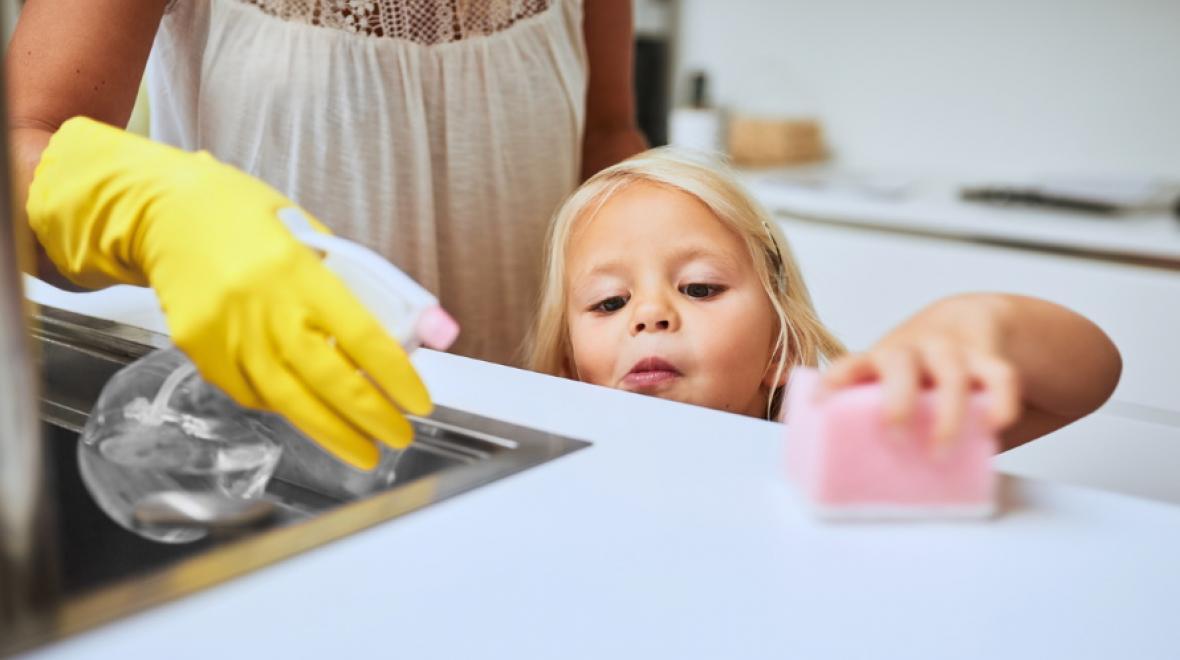 little girl on her tip-toes wiping the kitchen counter with a pink sponge