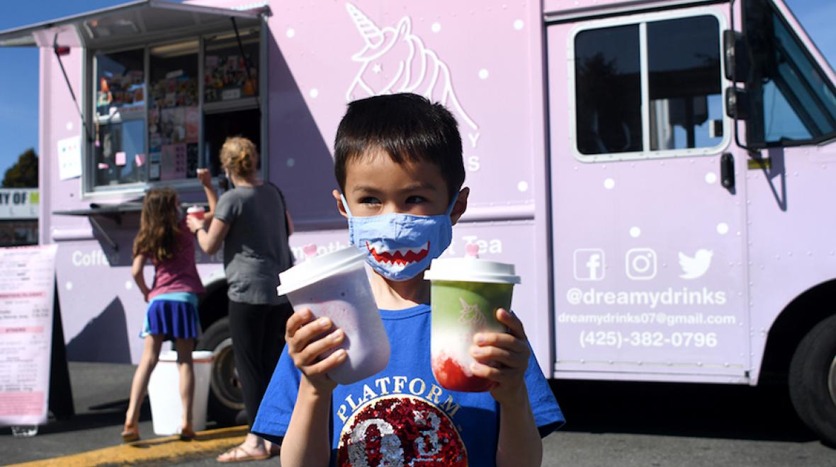 little boy with a shark mask holding two dreamy drinks with the dreamy drinks pink truck in the background