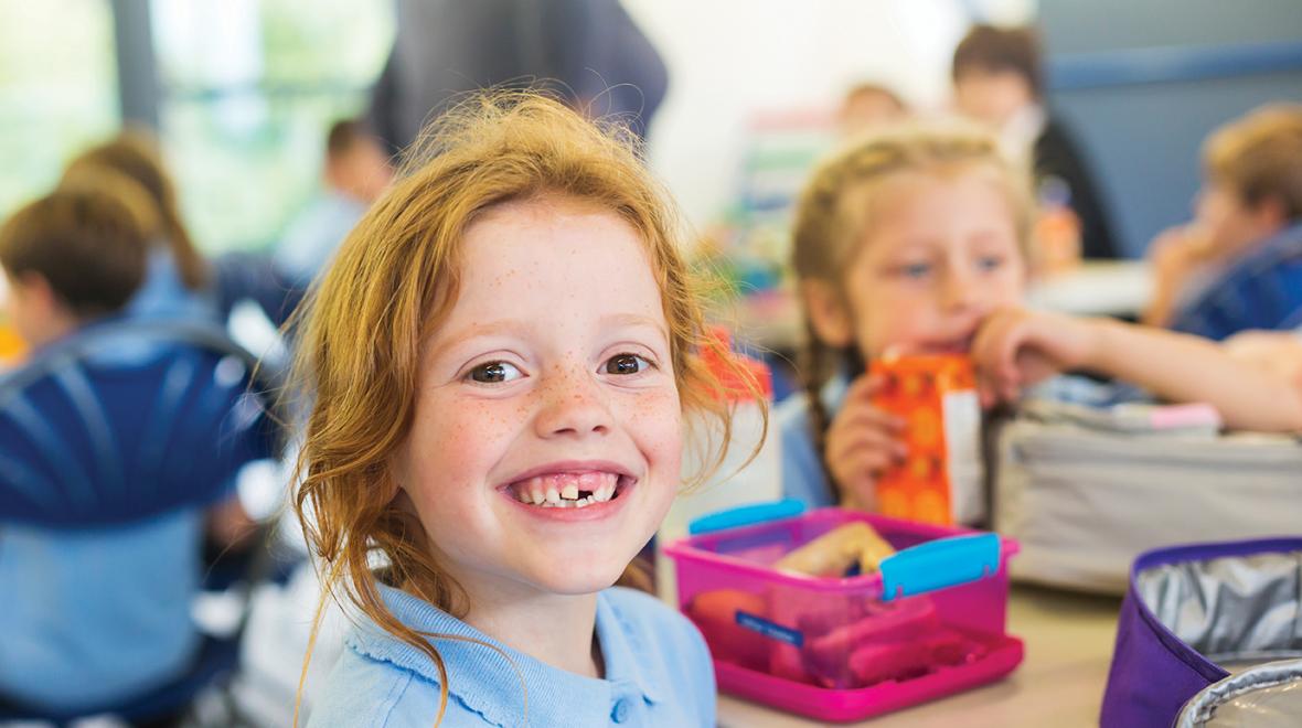 smiling redheaded girl sitting at a lunch table at school with lunchboxes and kids in the background