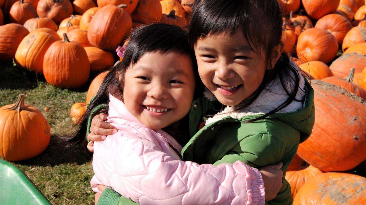 Two girls sisters siblings hugging in the pumpkin patch and posing for a photo. they're wearing jackets and the sun is shining