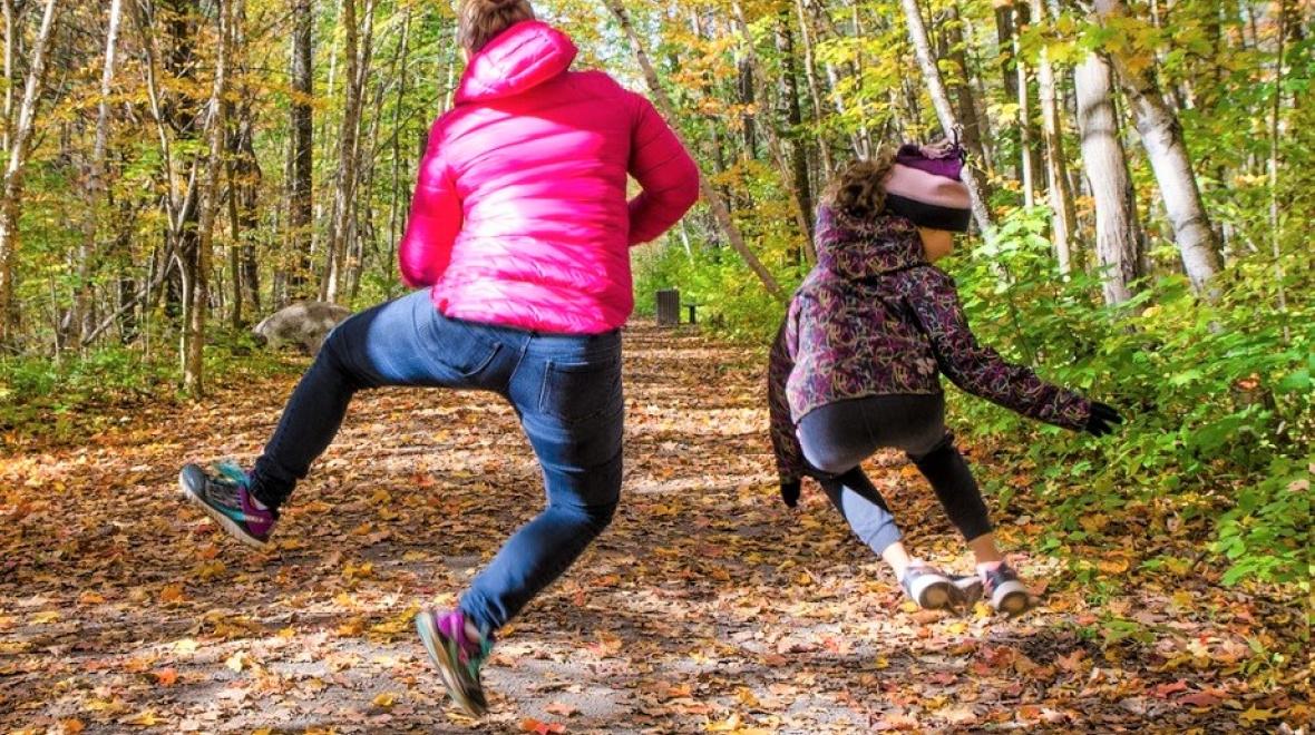 Mom and daughter jumping on a trail during a fall walk in the woods. They are wearing jackets and facing away from the camera