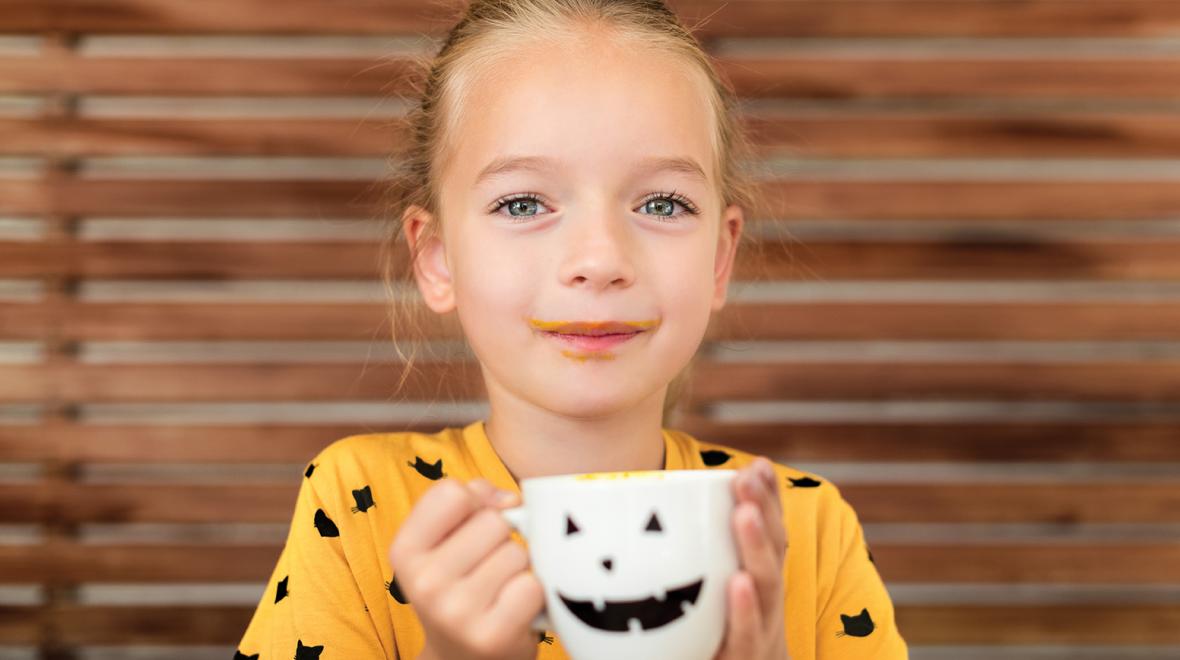 girl in an orange halloween shirt drinking out of a white jack o'lantern mug