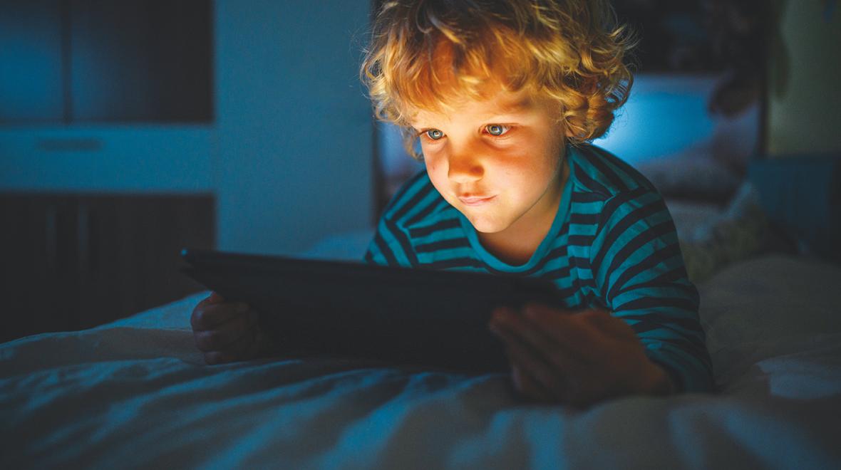 curly-haired blond boy laying on his bed in the dark with an ipad lighting up his face