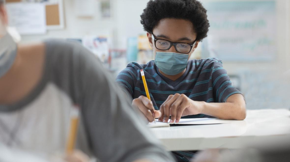 boy wearing glasses and a mask writing with a pencil at his desk in a school classroom