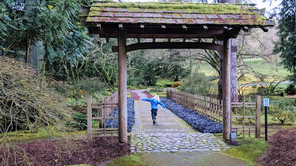 A young girl in a blue jacket has her arms outstretched as she walks a path at Bloedel Reserve a nature preserve on Bainbridge Island near Seattle
