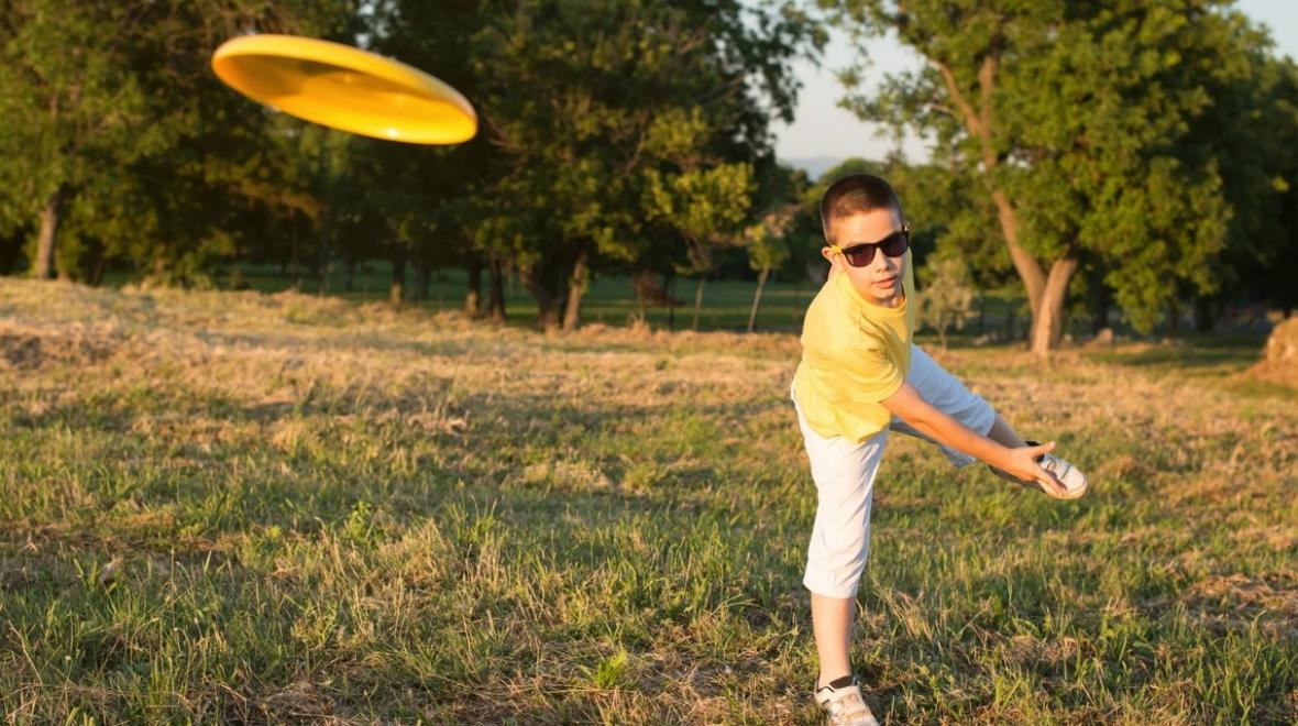 A school-age boy in a yellow shirt and sunglasses throws a yellow Frisbee disc during a game of disc golf free fun family sport to play in parks around Seattle