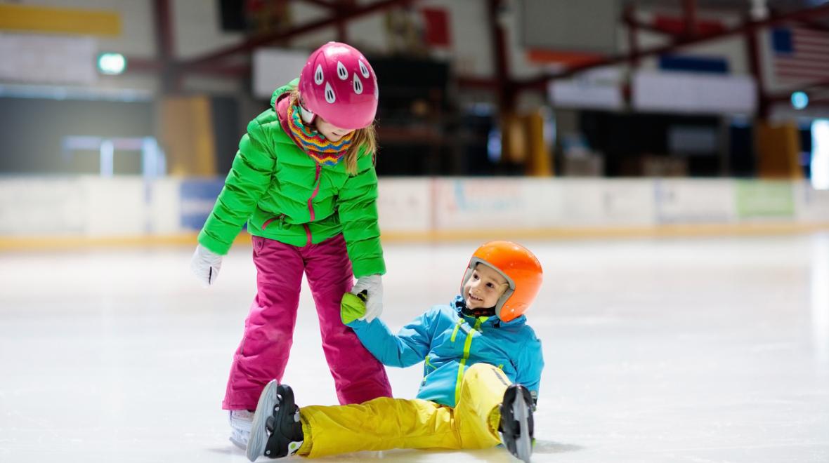 Kids in colorful winter clothes and wearing helmets ice skate at an indoor ice rink one has fallen and the other is helping her up