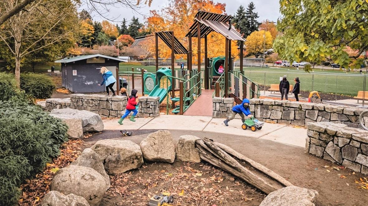 Kids play at updated Lakewood Playground in South Seattle. There's a sand pit in the foreground and a new play structure in the background and fall foliage viewable as well