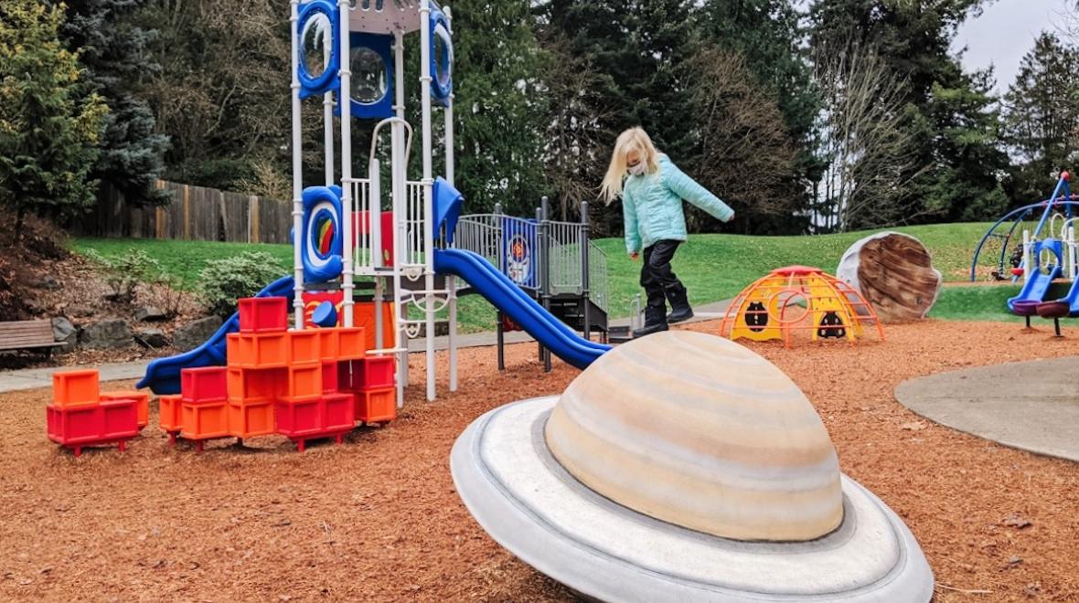 A girl stands on top of a planet-shaped play element at the newer playground at North Kirkland Community Center near Seattle among the best new playgrounds of 2021 winter break activity for kdis