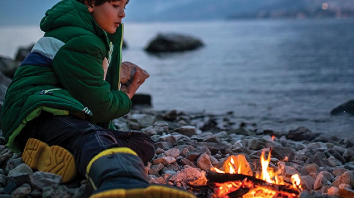 A young camper sits beside a fire on the shores of a lake