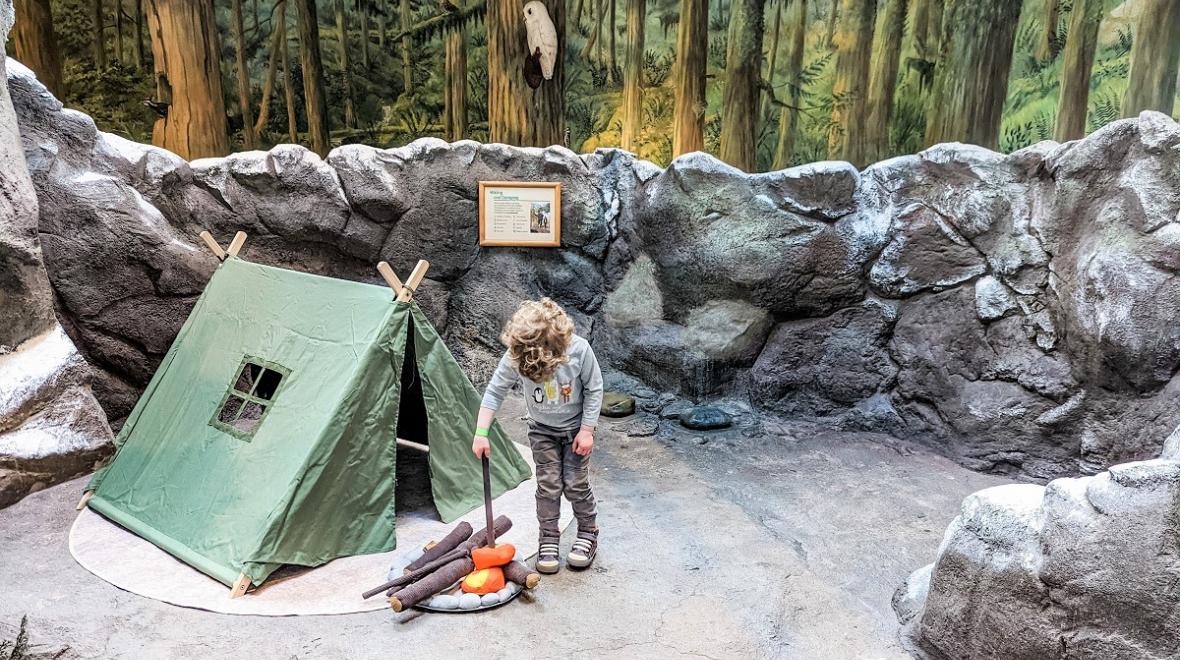 Young boy with curly hair tends a play campfire near a play camping tent on "the mountain" exhibit at Seattle Children's Museum reopening after a two-plus-year pandemic closure