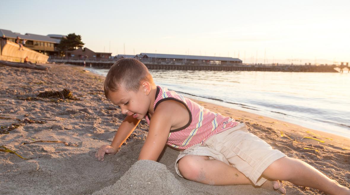 A young boy in shorts and a tank top plays in the sand on the beach in Edmonds, Washington. It's a sunny evening and a pier is visible behind him.