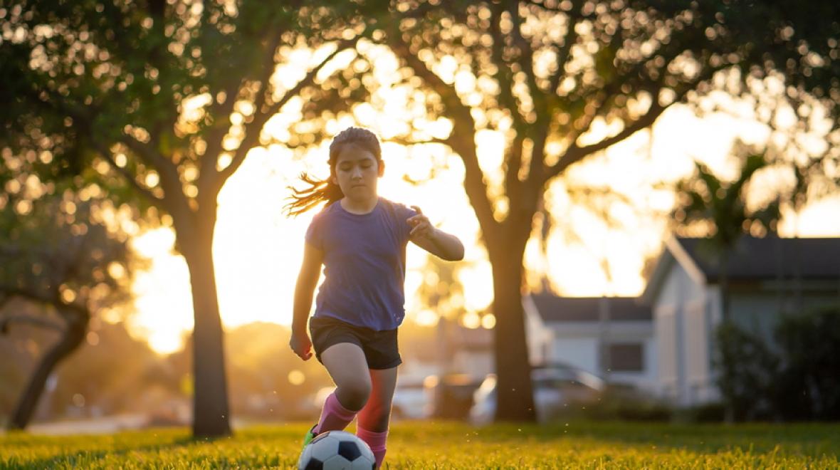 Young girl kicking a soccer ball in a field with trees behind her