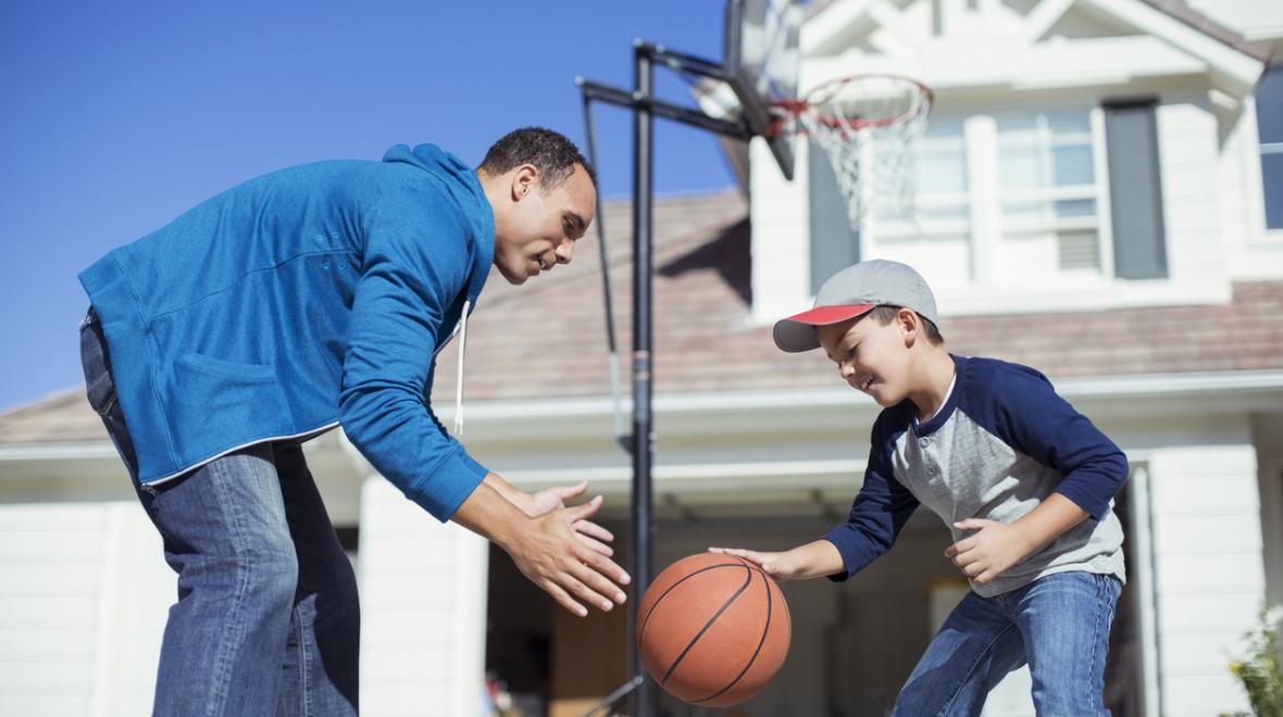 Father and son playing basketball outside of a big white house 