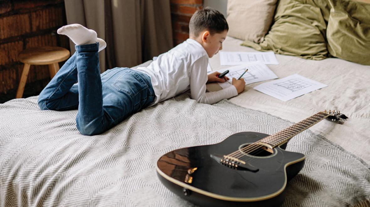 Young boy lying on his bed next to a guitar writing on a paper