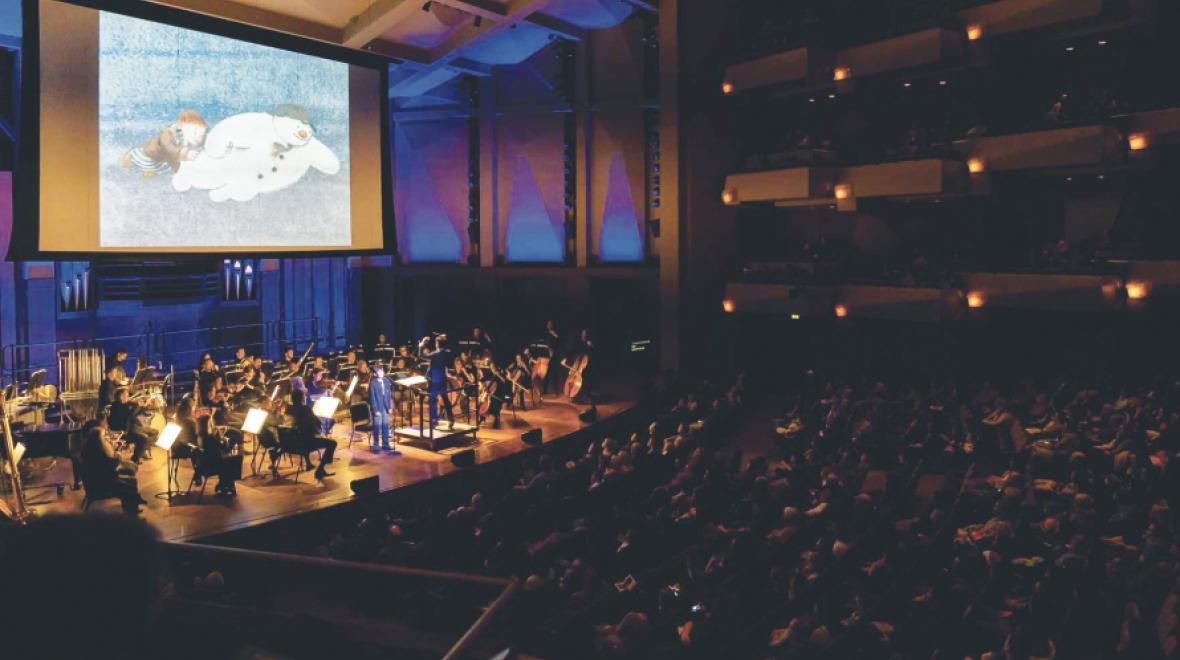 orchestra on a stage playing music with a video of a snowman playing behind them